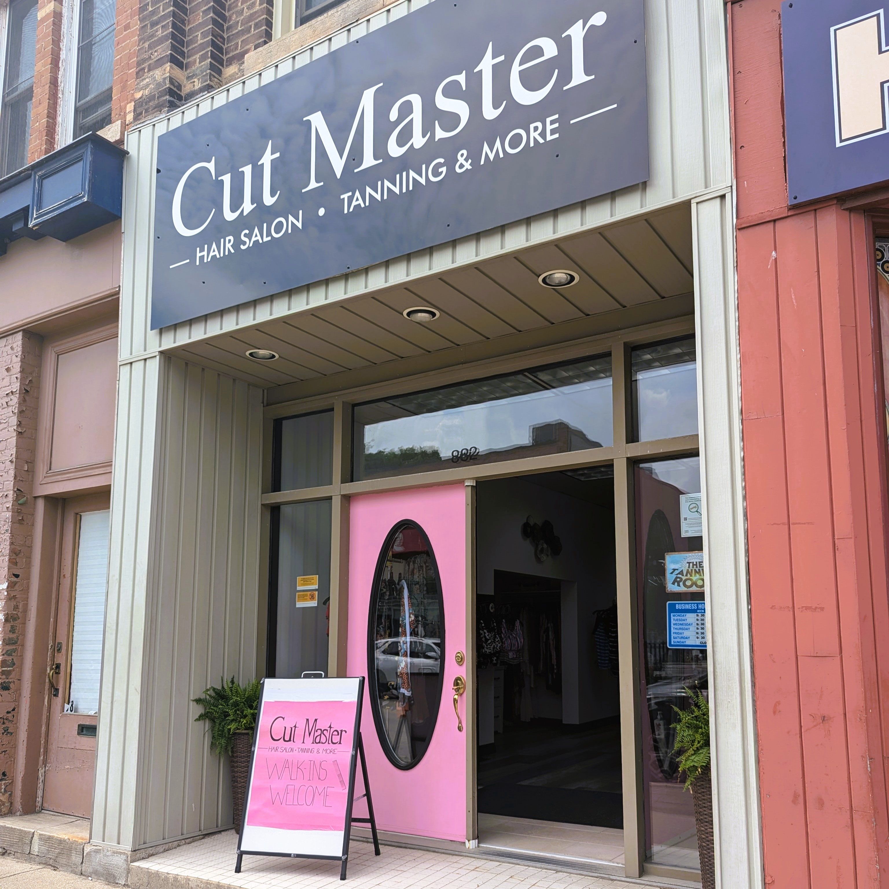 Double pink door with oval windows one door open to the inside of the building. A frame sign on step infront of door. Large black sign with business name above the door frame